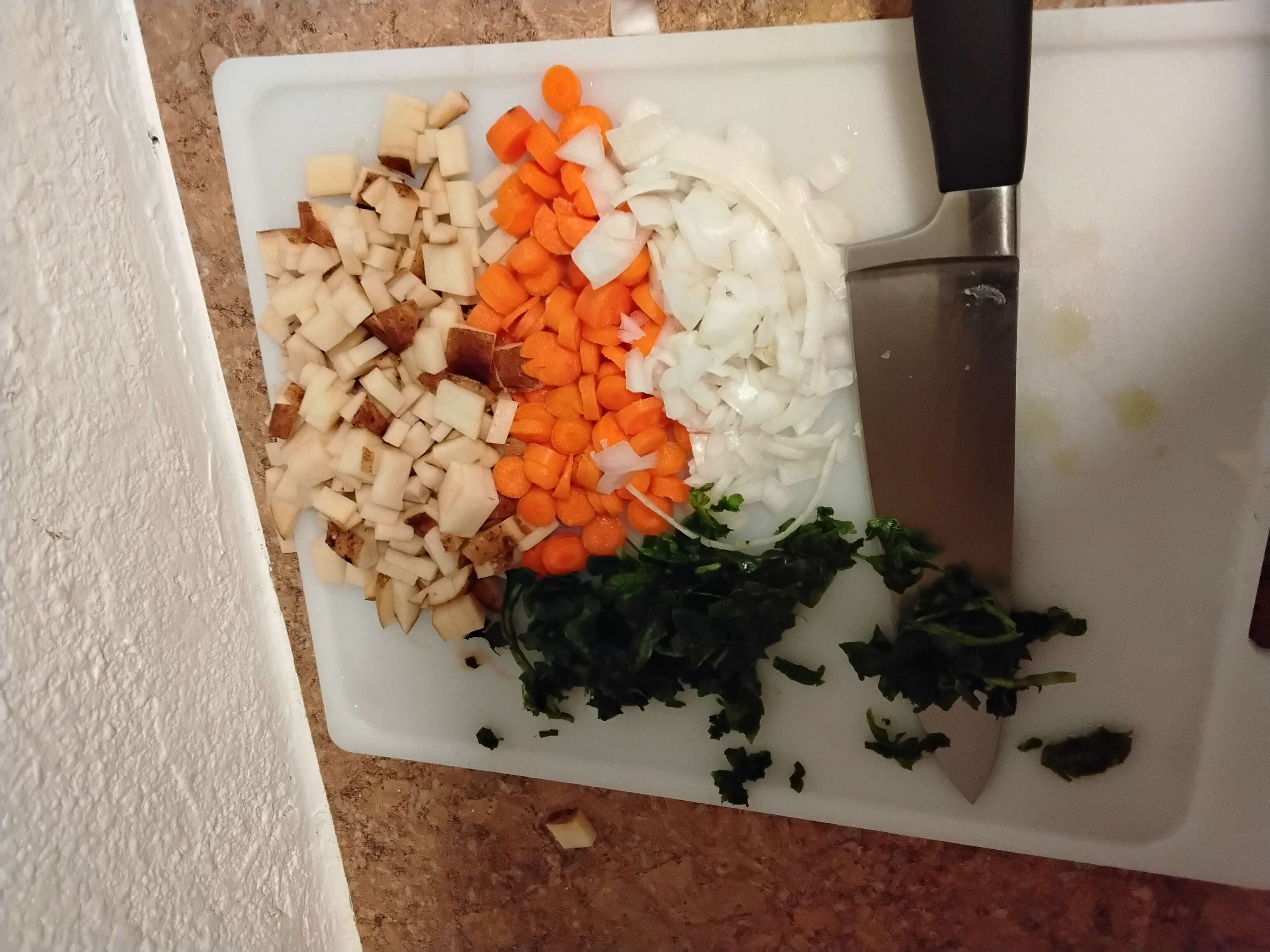 Mise en place: diced russet potato, sliced carrot rounds, diced white onion, and chiffonade spinach on a white cutting board with chef's knife